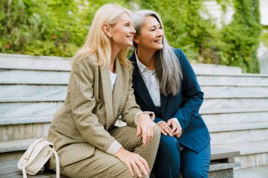 Olgun, çok kültürlü iş kadınları bankta otururken konuşuyor ve gülümsüyor.