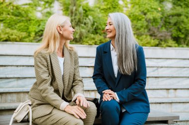 Olgun, çok kültürlü iş kadınları bankta otururken konuşuyor ve gülümsüyor.