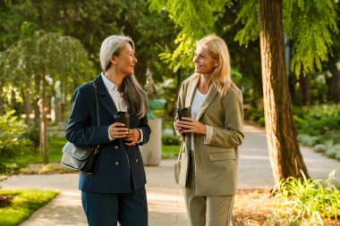 Olgun, çok kültürlü iş kadınları parkta yürürken kahve içiyorlar.