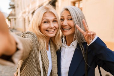 Şehir caddesinde selfie çekerken el kol hareketi yapan olgun kadınlar.