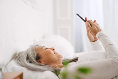 Gri Asyalı kadın evde uyuduktan sonra yatarken cep telefonu kullanıyor.