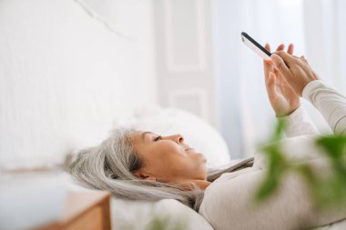 Gri Asyalı kadın evde uyuduktan sonra yatarken cep telefonu kullanıyor.