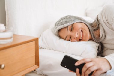 Gri Asyalı kadın evde uyuduktan sonra yatarken cep telefonu kullanıyor.