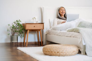 Gri Asyalı kadın evde uyuduktan sonra yatağında oturup gülerken ve dizüstü bilgisayar kullanırken