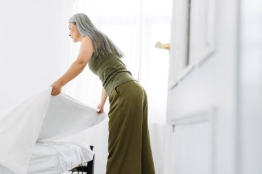 Gri saçlı, olgun Asyalı kadın sabah yatağını yapıyor.