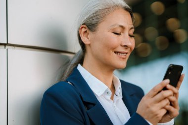 Dışarıda cep telefonu kullanırken gülümseyen gri Asyalı kadın.