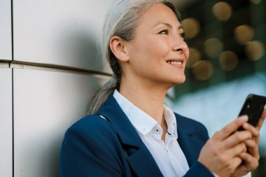 Dışarıda cep telefonu kullanırken gülümseyen gri Asyalı kadın.