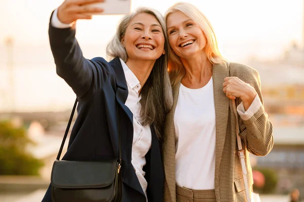 Olgun çok ırklı iş kadınları açık havada buluşurken cep telefonlarıyla selfie çekiyorlar.