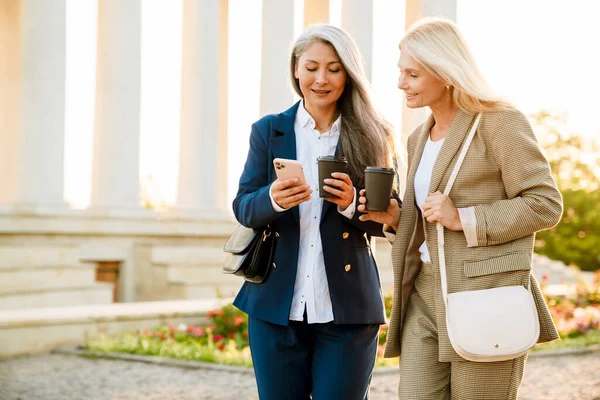 Olgun, çok ırklı iş kadınları dışarıda buluşurken cep telefonu kullanıyor ve kahve içiyor.