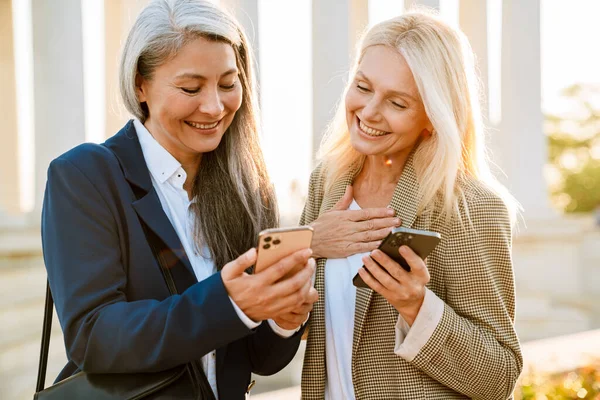 Açık havada buluşurken gülümseyen ve cep telefonu kullanan çok kültürlü iş kadınları.