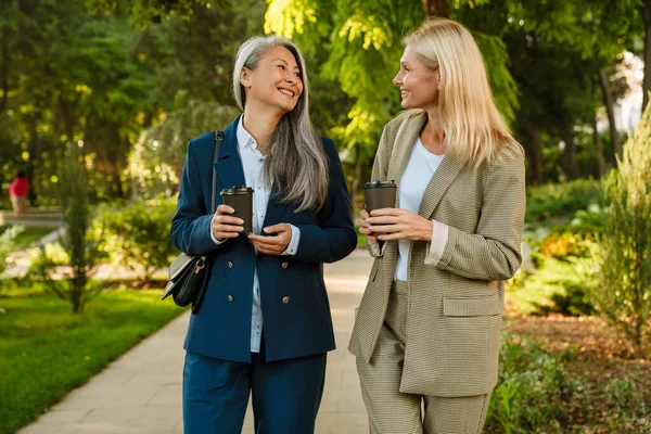 Olgun, çok kültürlü iş kadınları parkta yürürken kahve içiyorlar.