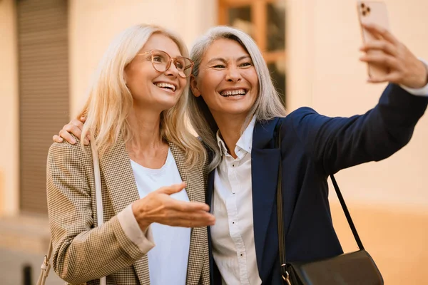 Şehirli caddede cep telefonuyla selfie çekerken gülümseyen olgun çok ırklı kadınlar.