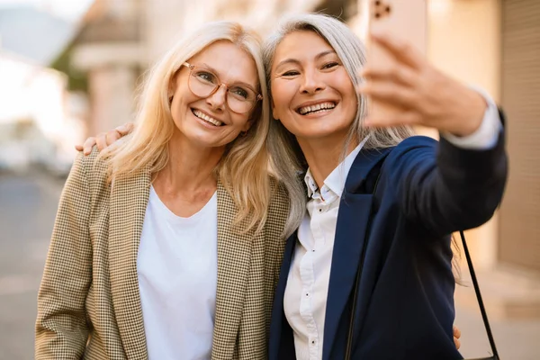 Şehirli caddede cep telefonuyla selfie çekerken gülümseyen olgun çok ırklı kadınlar.