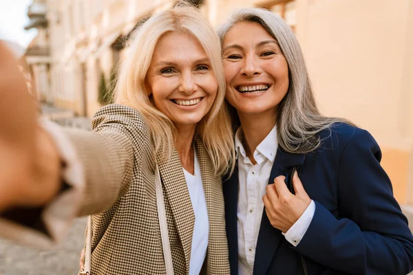 Şehir caddesinde selfie çekerken gülümseyen olgun çok ırklı kadınlar.