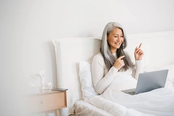 Grey asian woman pointing fingers aside and using laptop while sitting in bed at home