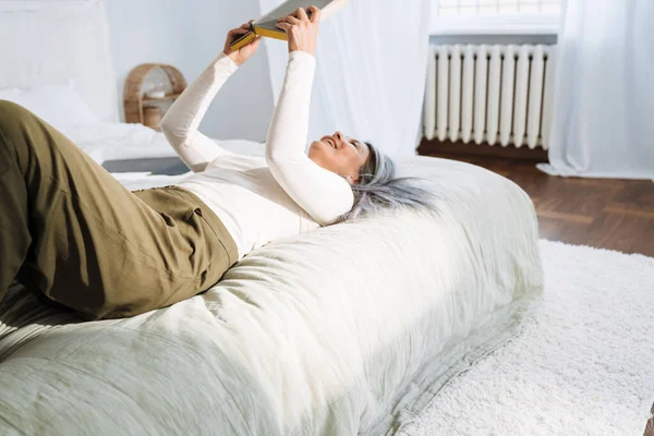 灰色アジアの女性笑顔と読書本ながら横たわっオンベッドに家 — ストック写真