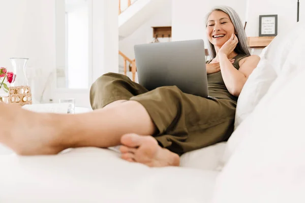 Mature Asian Woman Laughing Working Laptop While Sitting Couch Home — Foto de Stock