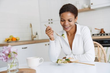 Genç siyahi kadın mutfakta yemek yerken gülümsüyor.