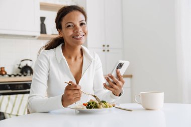 Genç siyahi kadın mutfakta yemek yerken cep telefonu kullanıyor.