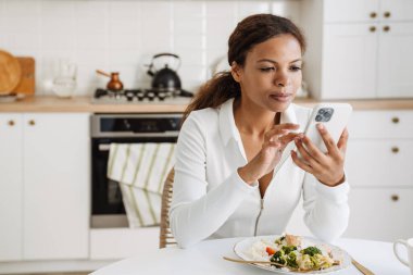 Genç siyahi kadın mutfakta yemek yerken cep telefonu kullanıyor.