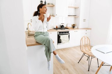 Genç siyah kadın mutfakta kahve içerken gülüyor.