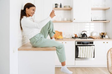 Mutfakta cep telefonu kullanırken gülümseyen genç siyahi kadın.