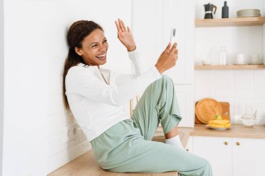 Genç siyahi kadın mutfakta cep telefonu kullanırken el kol hareketi yapıyor.