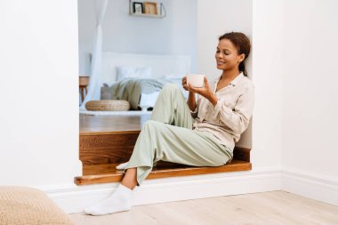 Siyah kadın evde merdivenlerde oturup çay içerken gülümsüyor.