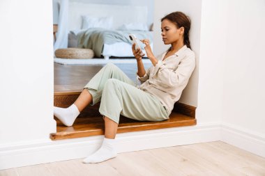 Genç siyahi kadın evdeki merdivenlerde otururken cep telefonu kullanıyor.