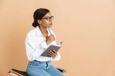 Genç siyahi kadın bej arka planda izole edilmiş bir sandalyede otururken notlar yazıyor.