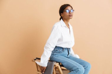Gözlüklü genç siyahi kadın bej arka planda izole edilmiş bir sandalyede oturuyor.