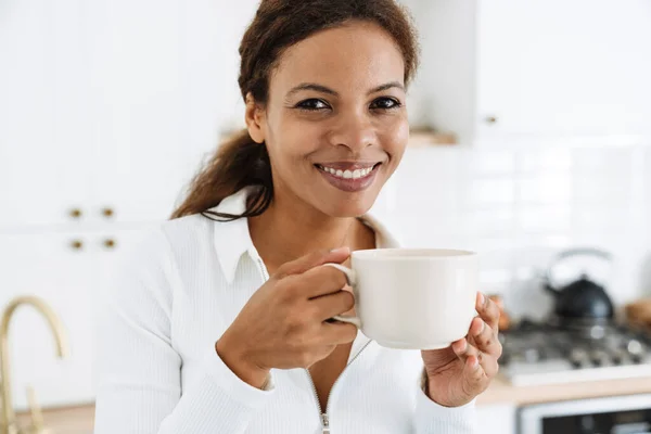 家のキッチンでコーヒーを飲みながら笑顔の若い黒人女性 — ストック写真