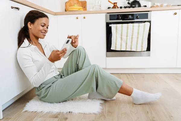 年轻的黑人妇女坐在厨房的地板上 一边用手机 — 图库照片