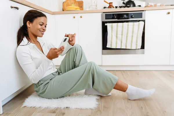 Young Black Woman Using Cellphone While Sitting Floor Kitchen Home — стоковое фото