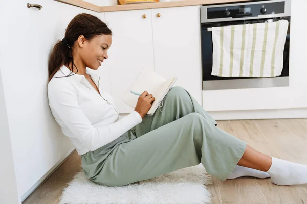 Young Black Woman Writing Notes While Sitting Floor Kitchen Home — стоковое фото