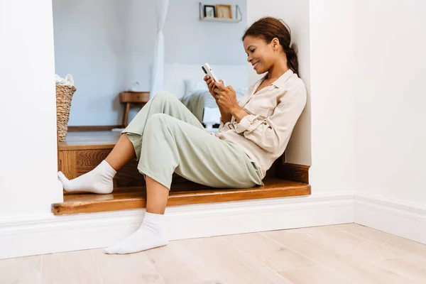 年轻的黑人妇女坐在楼梯上用手机在家里 — 图库照片