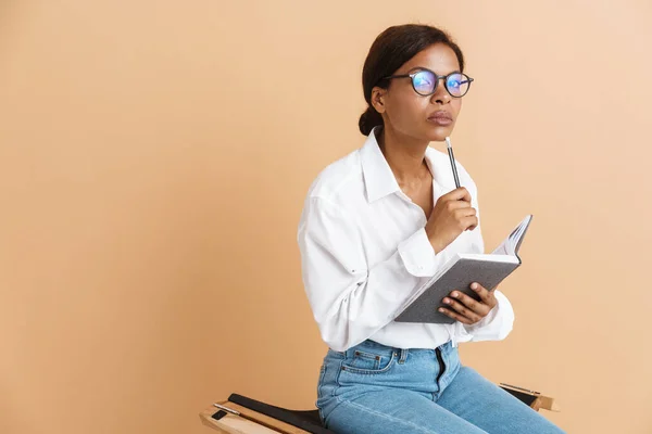 Young black woman writing down notes while sitting on chair isolated over beige background