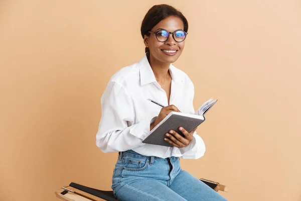 Young black woman writing down notes while sitting on chair isolated over beige background