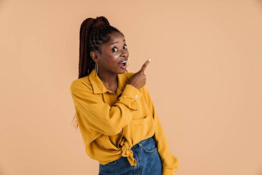 Genç siyahi kadın şaşkınlığını dile getiriyor ve bej arka planda izole edilmiş bir şekilde işaret ediyor.