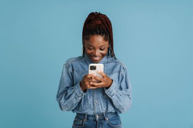 Young black woman with afro pigtails smiling and using cellphone isolated over blue background