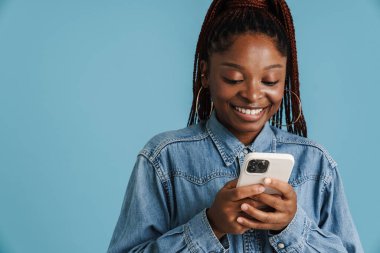 Siyah saçlı, saçları örgülü, gülümseyen ve cep telefonunu mavi arka planda kullanan genç bir kadın.