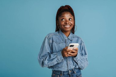 Siyah saçlı, saçları örgülü, gülümseyen ve cep telefonunu mavi arka planda kullanan genç bir kadın.