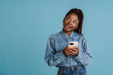 Mavi arka planda izole edilmiş cep telefonu kullanan genç siyahi bir kadın dilini ısırıyor.