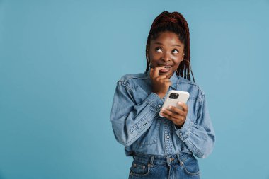 Mavi arka planda izole edilmiş cep telefonu kullanan genç siyahi bir kadın parmağını ısırıyor.
