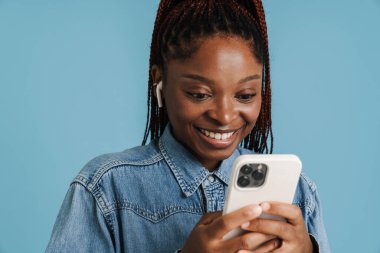 Mavi arka planda izole edilmiş cep telefonu ve kulaklık kullanırken gülümseyen genç siyahi kadın.