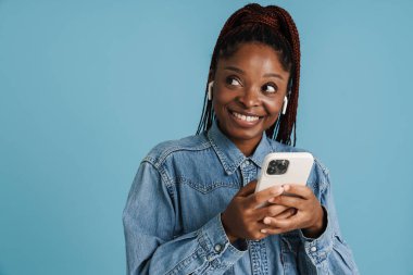 Mavi arka planda izole edilmiş cep telefonu ve kulaklık kullanırken gülümseyen genç siyahi kadın.