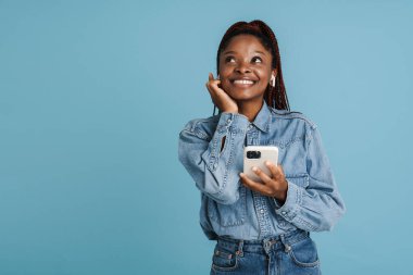 Mavi arka planda izole edilmiş cep telefonu ve kulaklık kullanırken gülümseyen genç siyahi kadın.