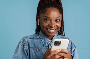 Mavi arka planda izole edilmiş cep telefonu ve kulaklık kullanırken gülümseyen genç siyahi kadın.