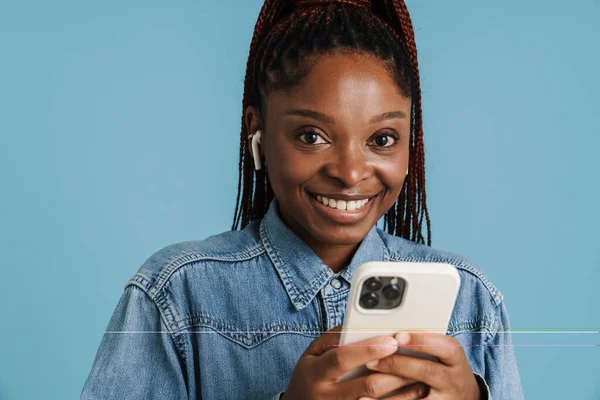 Mavi arka planda izole edilmiş cep telefonu ve kulaklık kullanırken gülümseyen genç siyahi kadın.