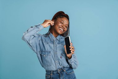 Mavi arka planda izole edilmiş müzik dinlerken gülümseyen ve dans eden genç siyahi kadın.